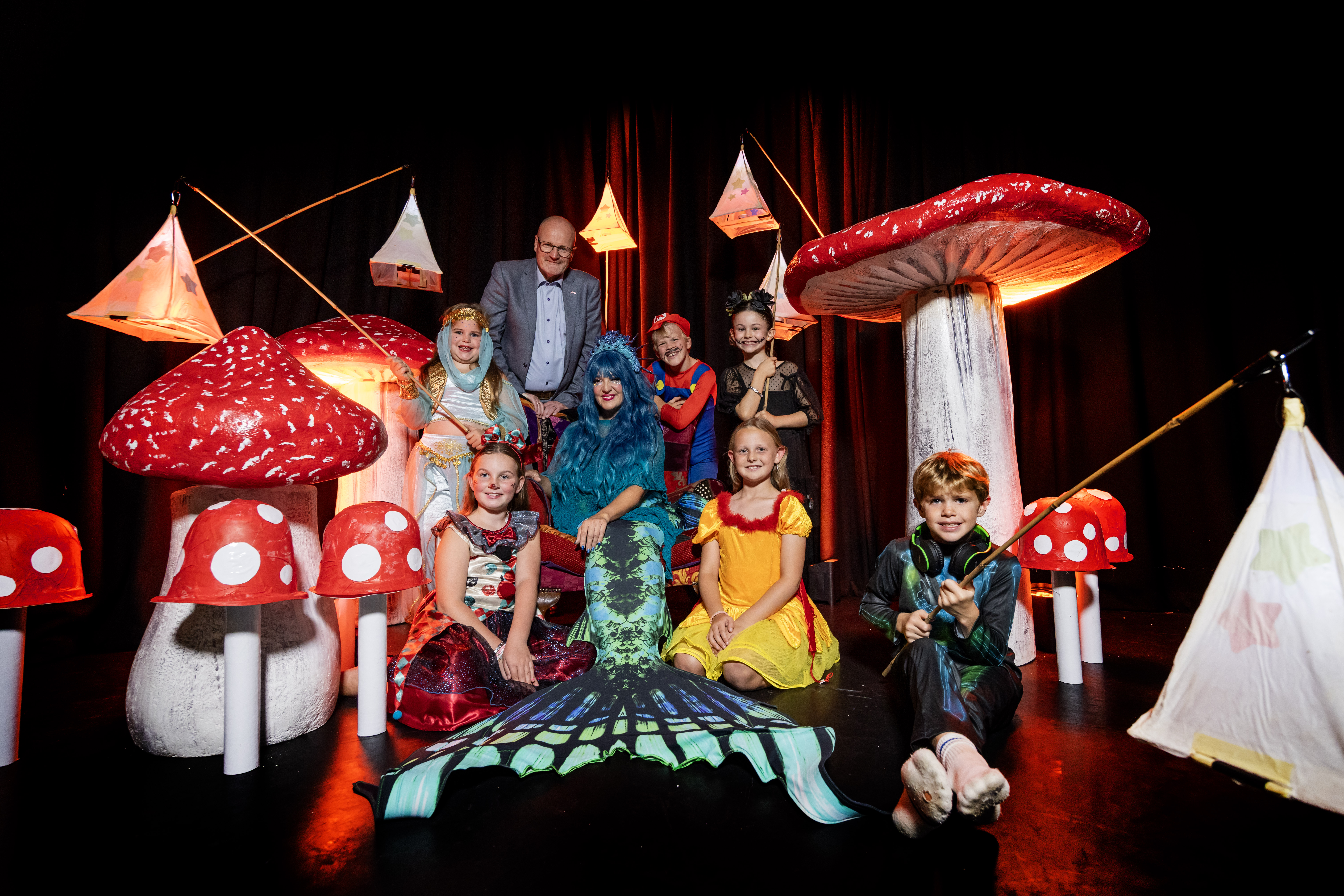 A group of performers and children in costumes inspired by fantasy are posing together on a stage. The set features large, whimsical toadstools and glowing lanterns. In the centre, an adult performer is seated, wearing a blue wig and a scaly, mermaid-like 