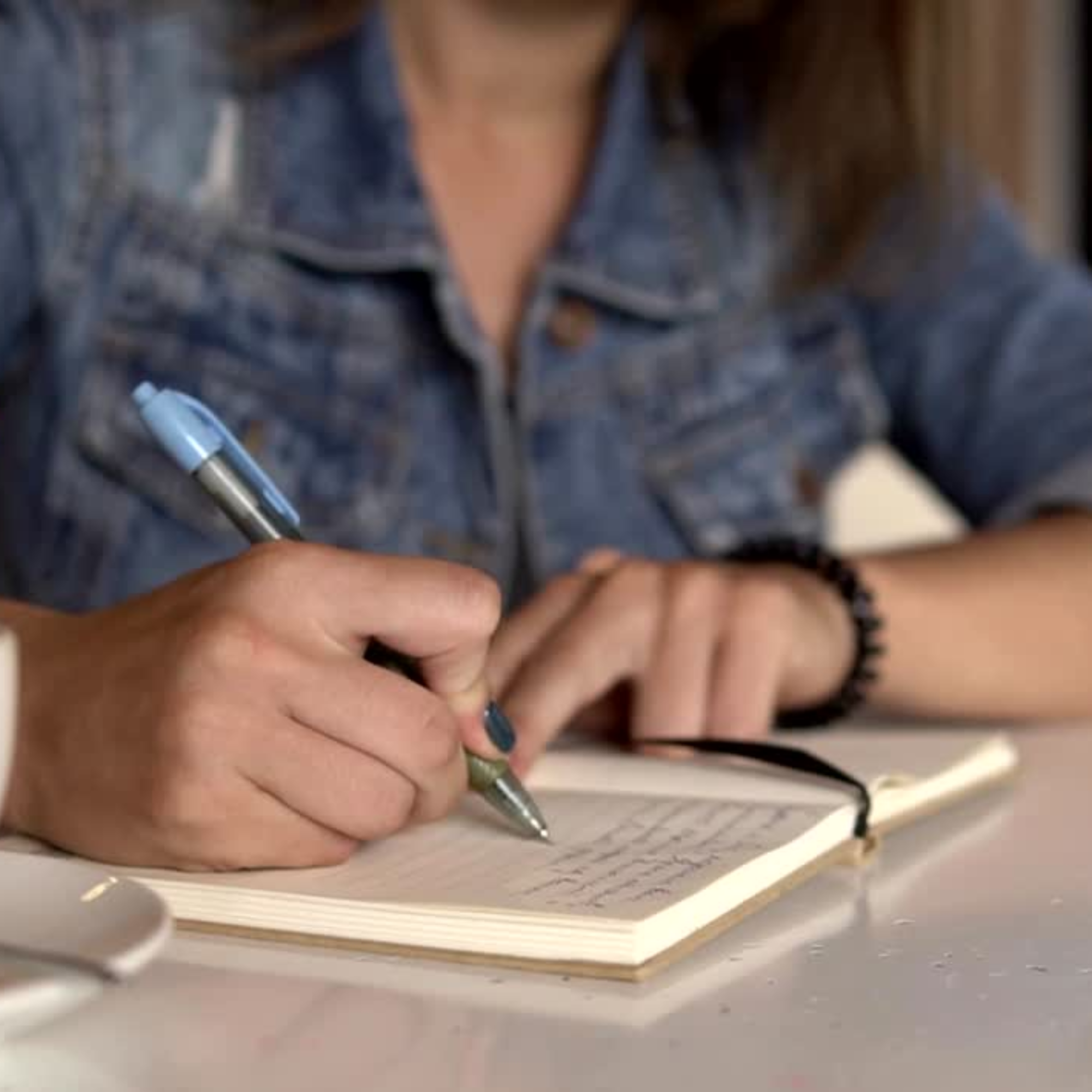  Close-up of a person wearing a denim jacket and a beaded bracelet, writing in a small open notebook with a blue pen. Their fingers are visible, and they have dark nail polish. The notebook sits on a bright, speckled white surface.