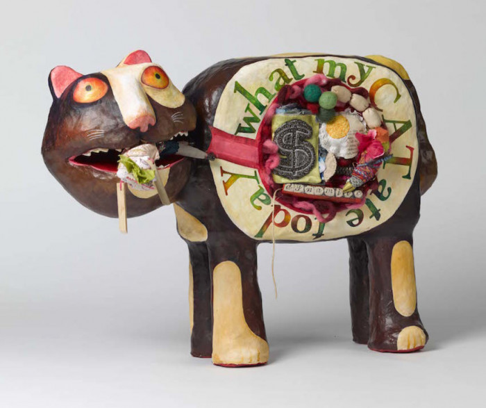 Mixed media artwork of found materials the shape of a 3d cat.  There is an open view of the cat's stomach and contents which are satirical in nature.