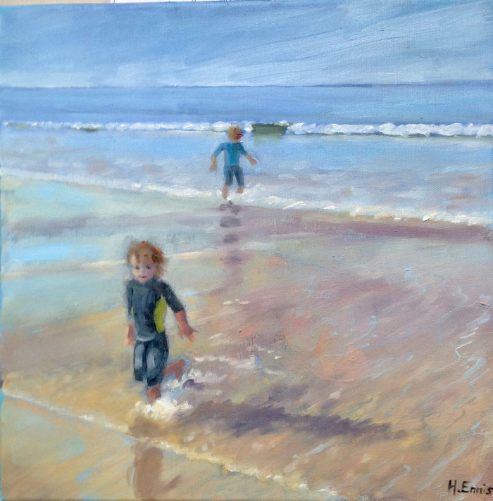 Two small children paddling at the water's edge at Portstewart Strand.  The sea is bluw with lapping waves on the yellow sandy shore.  The children have ginger curly hair and navy jumpers and blue jeans.